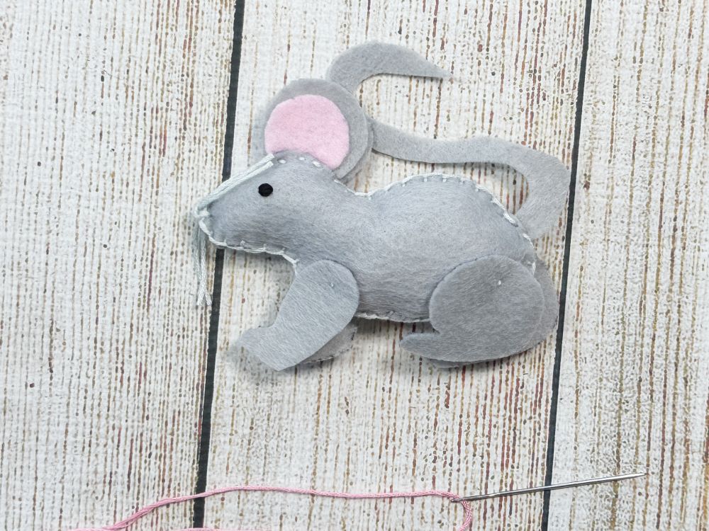 Finishing a felt mouse by hand stitching a pink nose with embroidery floss.