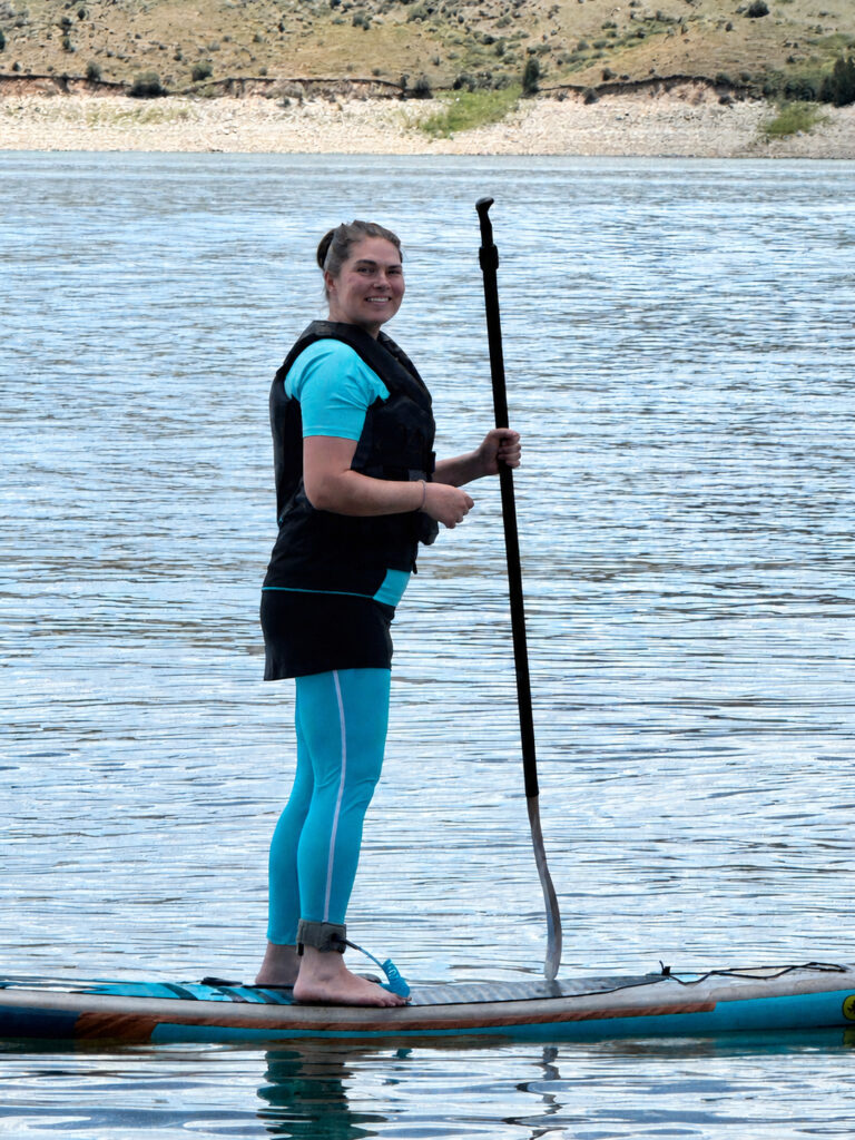 Woman wearing a turquoise Halcurt short sleeve rash guard while stand-up paddle boarding on a lake during summer.